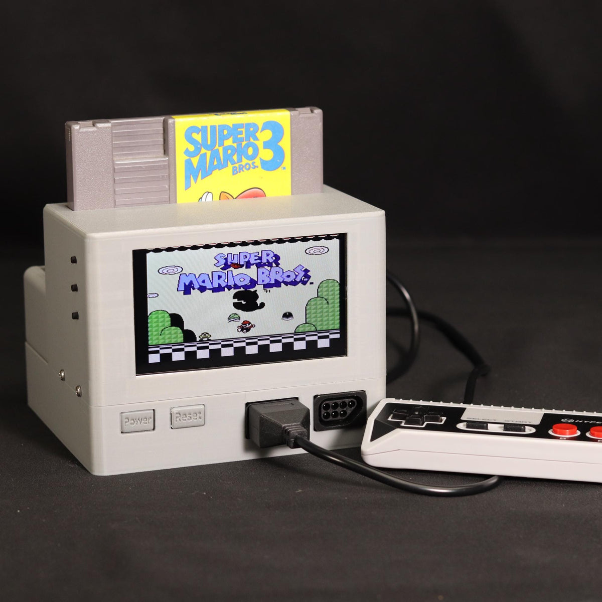 A retro-style console device with a Super Mario 3 cartridge inserted, a small screen showing the game, and a white controller with red buttons placed in front on a dark background.