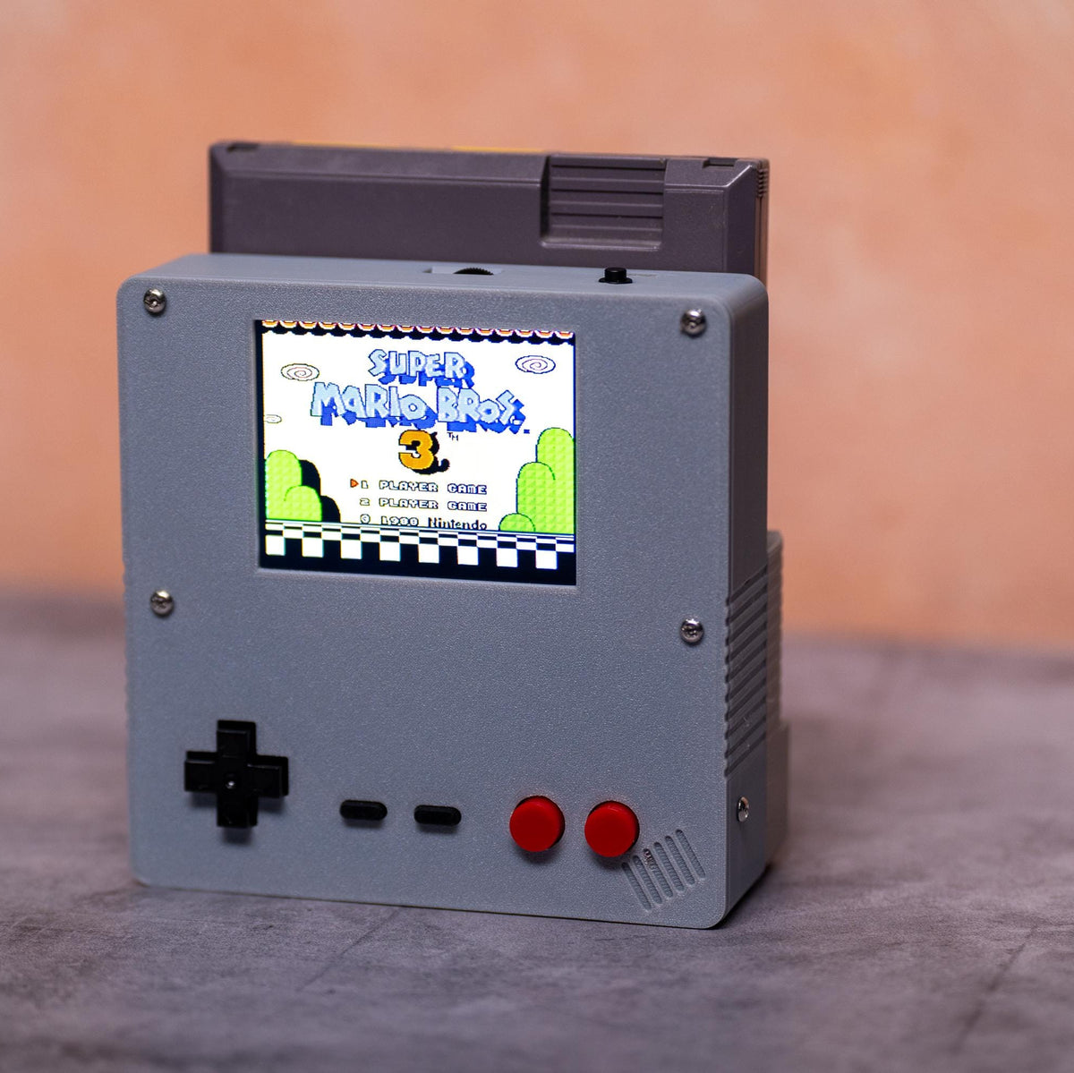 A retro-style gray handheld game console with a black D-pad, two red buttons, a small screen showing a colorful game menu, and a black cartridge docked on top, set against a warm peach background and gray surface.