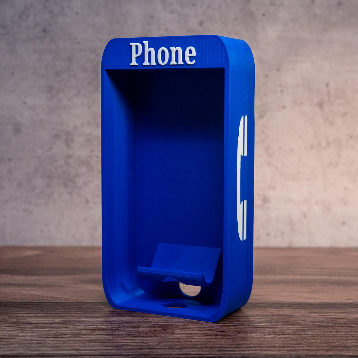 A bright blue phone-shaped object with the word 'Phone' printed in white at the top, standing on a wooden surface against a textured gray wall.
