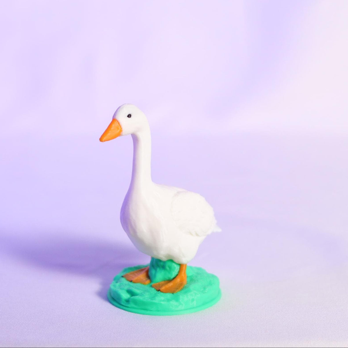 A small white goose figurine with an orange beak and orange feet, standing on a teal circular base against a soft lavender studio background.