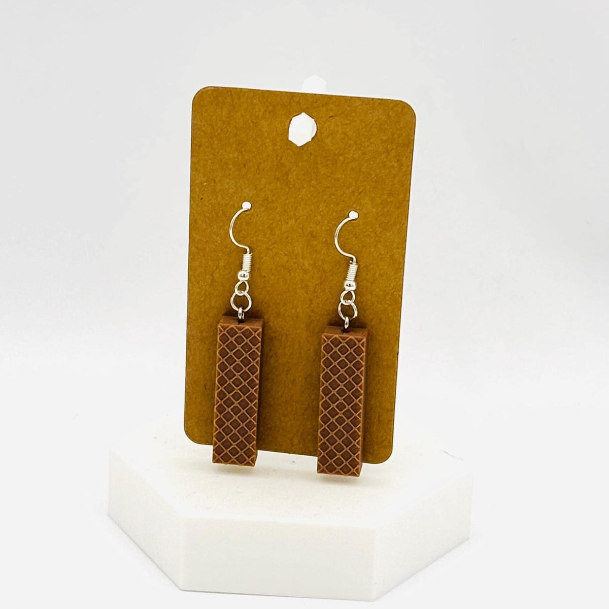 A pair of rectangular drop earrings with a brown and black textured pattern, hanging from silver fishhook wires, displayed on a brown cardboard card mounted on a white hexagonal stand against a white background.