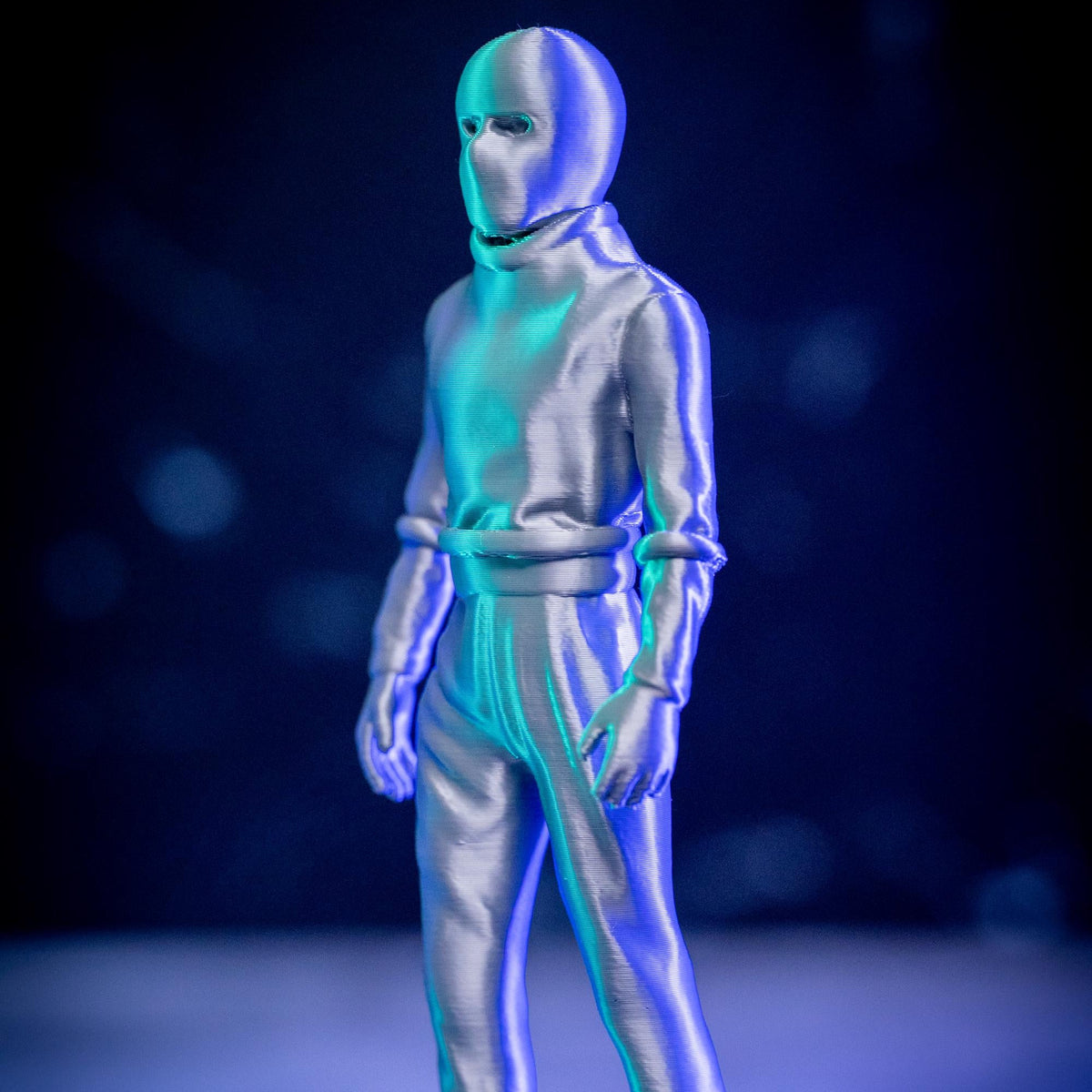 A full-body humanoid figure in a shiny metallic silver suit with a face mask, standing against a dark blue and black background with blue and cyan lighting reflections.