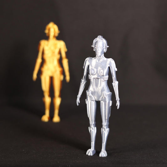 A metallic silver humanoid robot action figure stands in the foreground on a black background, with a blurred golden humanoid figure standing behind it.